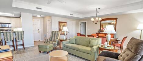 A spacious living room with a green sofa, a brown armchair, and a round coffee table. There is a kitchen area with a microwave and chairs.