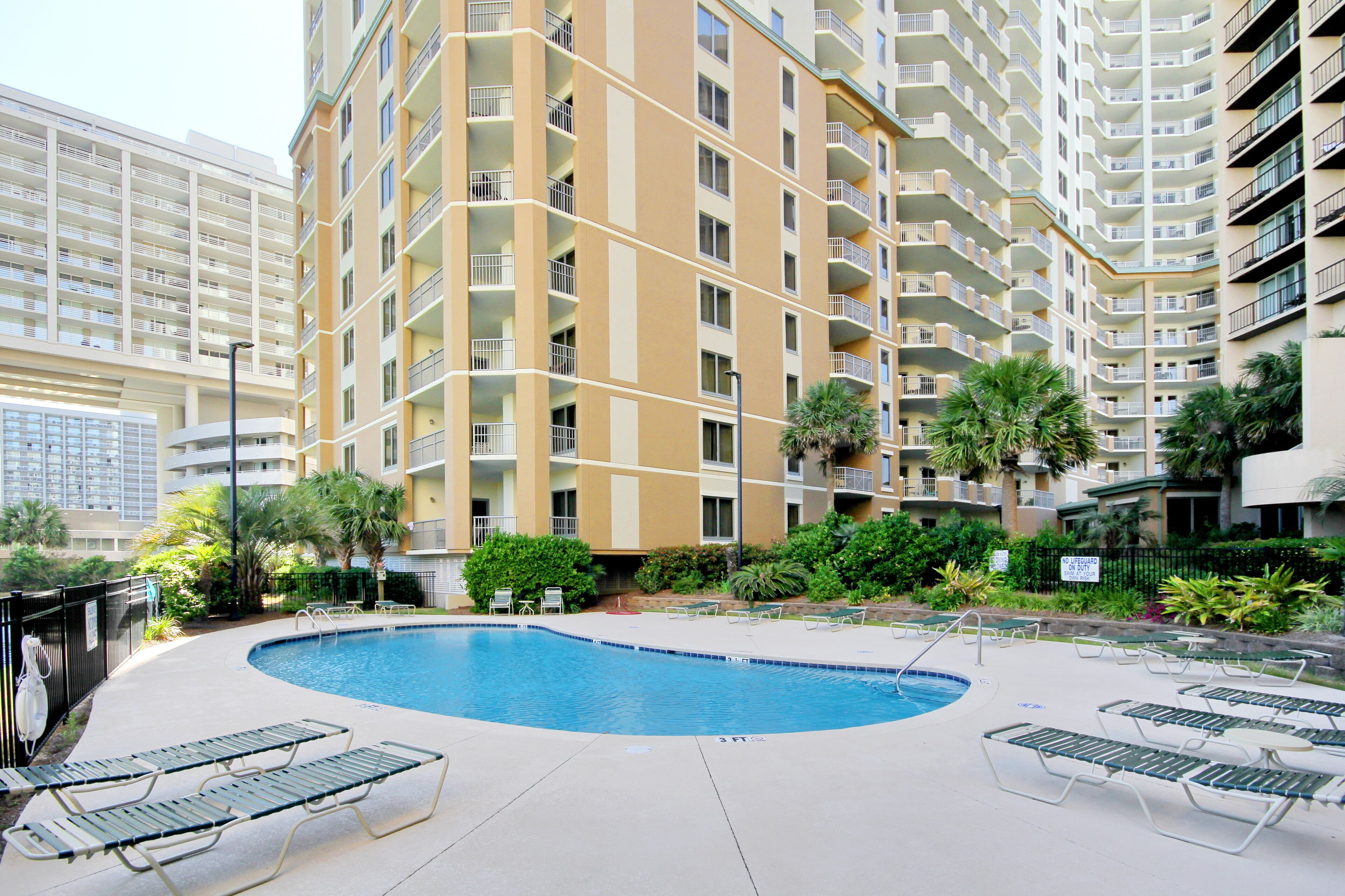 A hotel with a swimming pool surrounded by lounge chairs, palm trees, and a multi-story building with balconies.