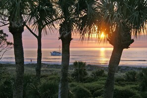 A sunset over a beach with palm trees and a boat in the distance.