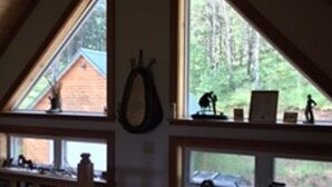 A cozy attic bedroom with a sloped ceiling, large windows offering a view of the outdoors, a bed with a checkered pillow, and a small desk area with a laptop and decorative items.