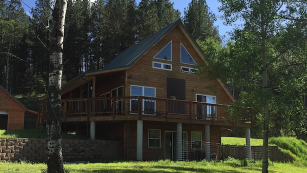 A two-story wooden cabin with a balcony, surrounded by a grassy yard and trees.