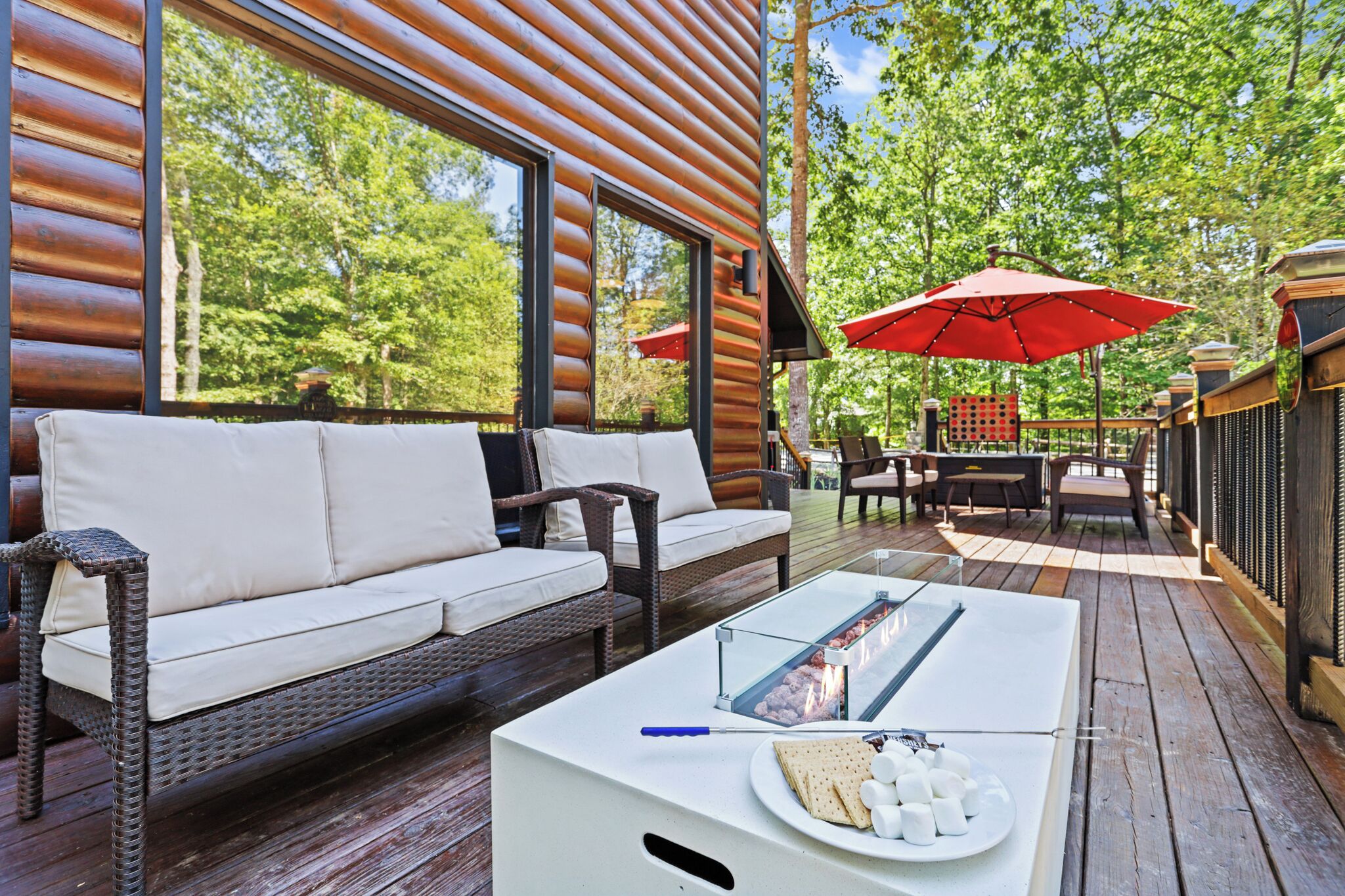 A wooden deck with a white sofa, a glass-top table with a fire feature, and outdoor furniture under red umbrellas.