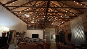 Amplia sala de estar con techo alto, vigas de madera a la vista y luces colgantes. Hay sofás de cuero, un televisor y una chimenea.