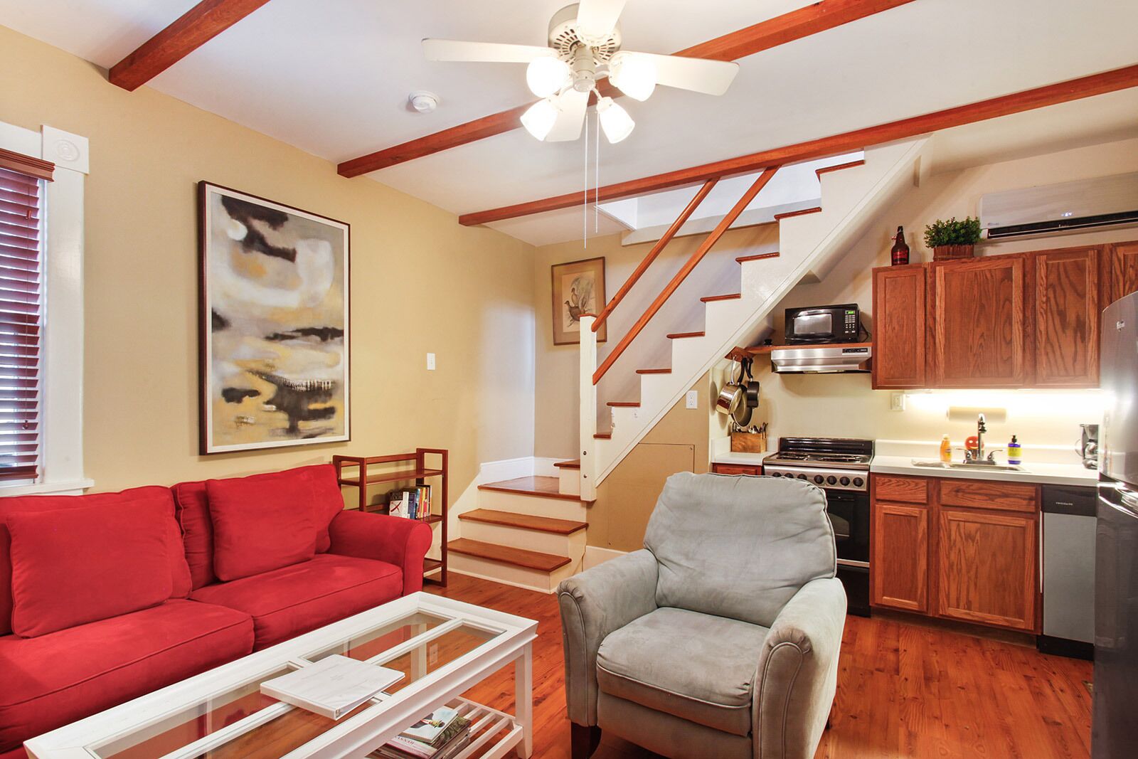A living room with a red sofa, a grey armchair, and a glass coffee table. A staircase leads to a kitchen with wooden cabinets and a microwave.
