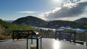 A table with a glass top and metal chairs, overlooking a coastal landscape with hills and houses. - Samba Azul 2 br2 ba ensuites w ACs private pool beautiful sunsets &new GENERATOR (St.John)