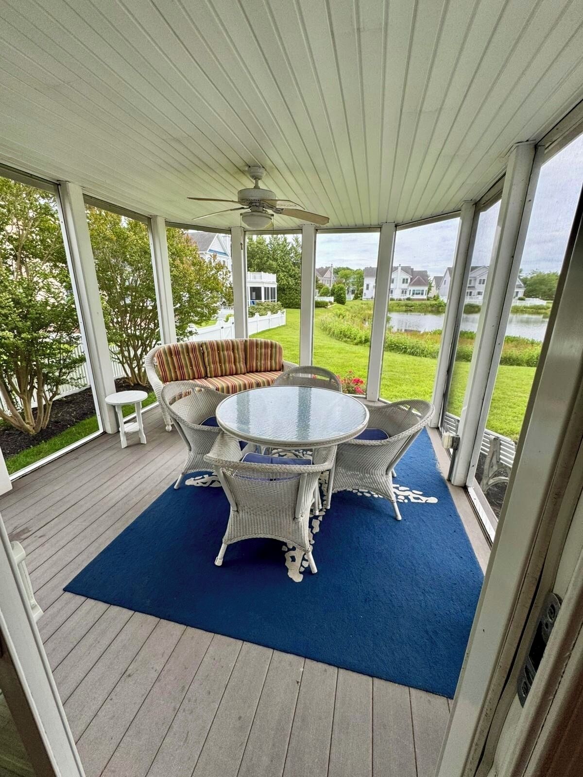 A covered patio with a round table and wicker chairs, a white bench, and a view of a lake and houses.