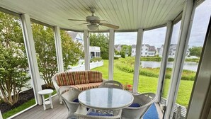A covered patio with a round table and wicker chairs, a white bench, and a view of a lake and houses. - Waterfront 5B+den Bethany Lakes, POOL,Pickleball,Gym, 5 min to beach, dogs ok (Bethany Beach)