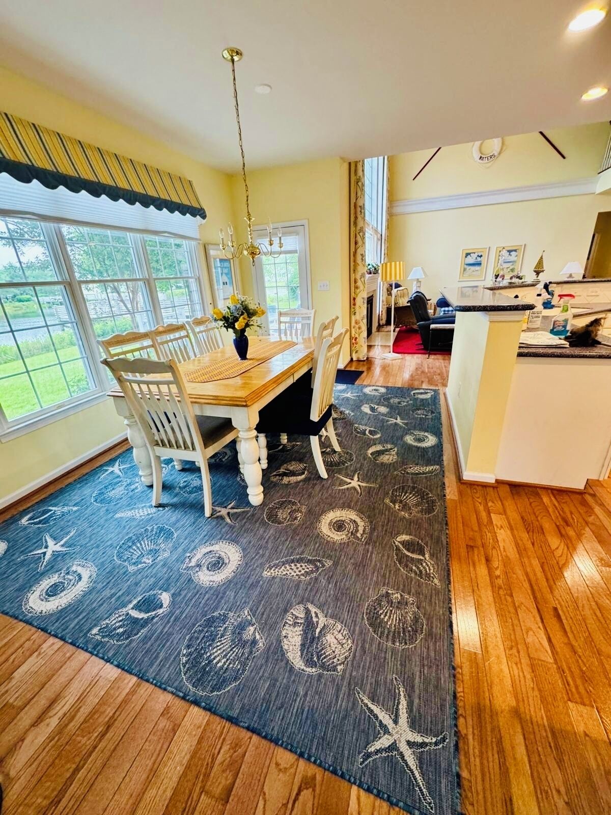 A dining area with a wooden table and chairs, a blue rug with sea shell patterns, and a view of the outdoors through large windows.