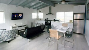 Eine moderne Küche mit weißen Schränken, einem eingebauten Ofen, einer Mikrowelle und einem Edelstahlkühlschrank. Es gibt einen Essbereich mit Tisch und Stühlen sowie ein Wohnzimmer mit einer Couch und einem an der Wand montierten Fernseher.