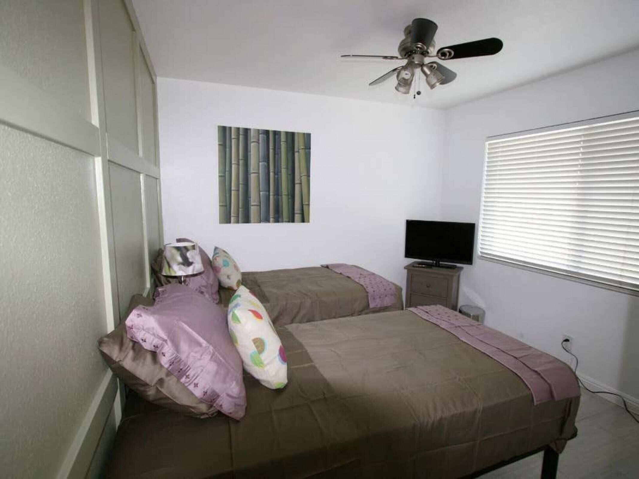 A bedroom with a bed, a ceiling fan, a television, and a window with blinds.