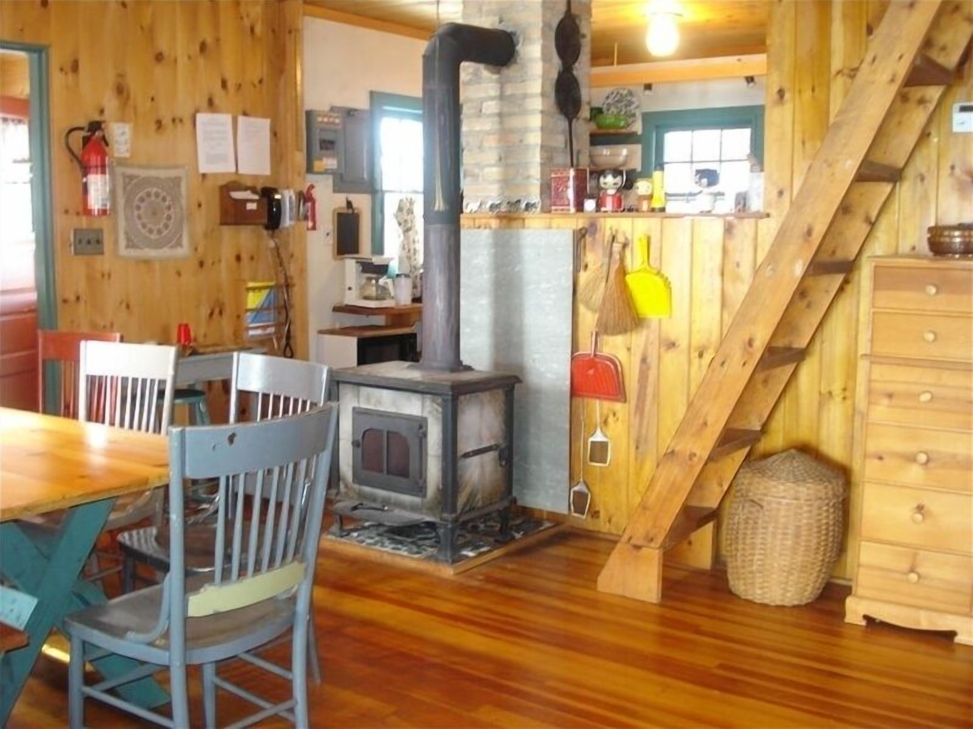 A rustic wooden interior with a central wood-burning stove, a dining area with chairs, and a staircase.