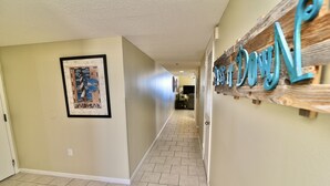 A hallway with a framed picture on the left, a wooden sign on the right, and a tiled floor.