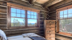 Un dormitorio tipo cabaña de madera con cama, cómoda y ventanas amplias que ofrecen vistas a árboles y montañas.