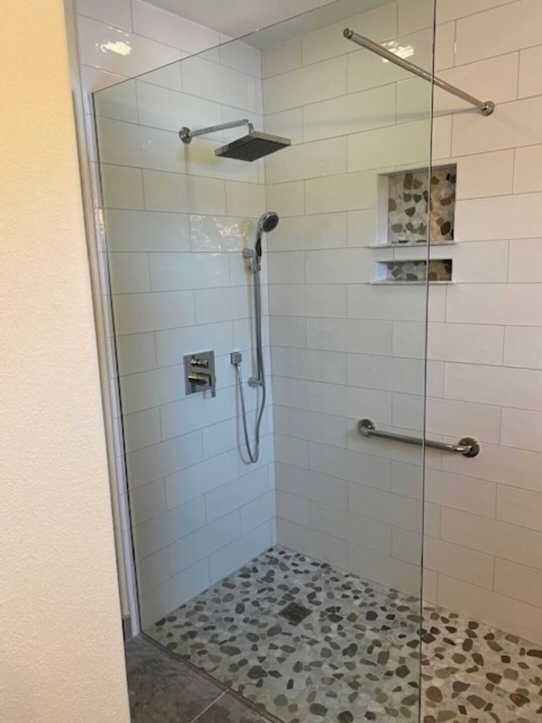 Une douche fermée par des parois en verre, dotée d’un pommeau de douche à main et d’un pommeau de douche fixe, avec des murs carrelés et un sol dont le motif rappelle des galets.