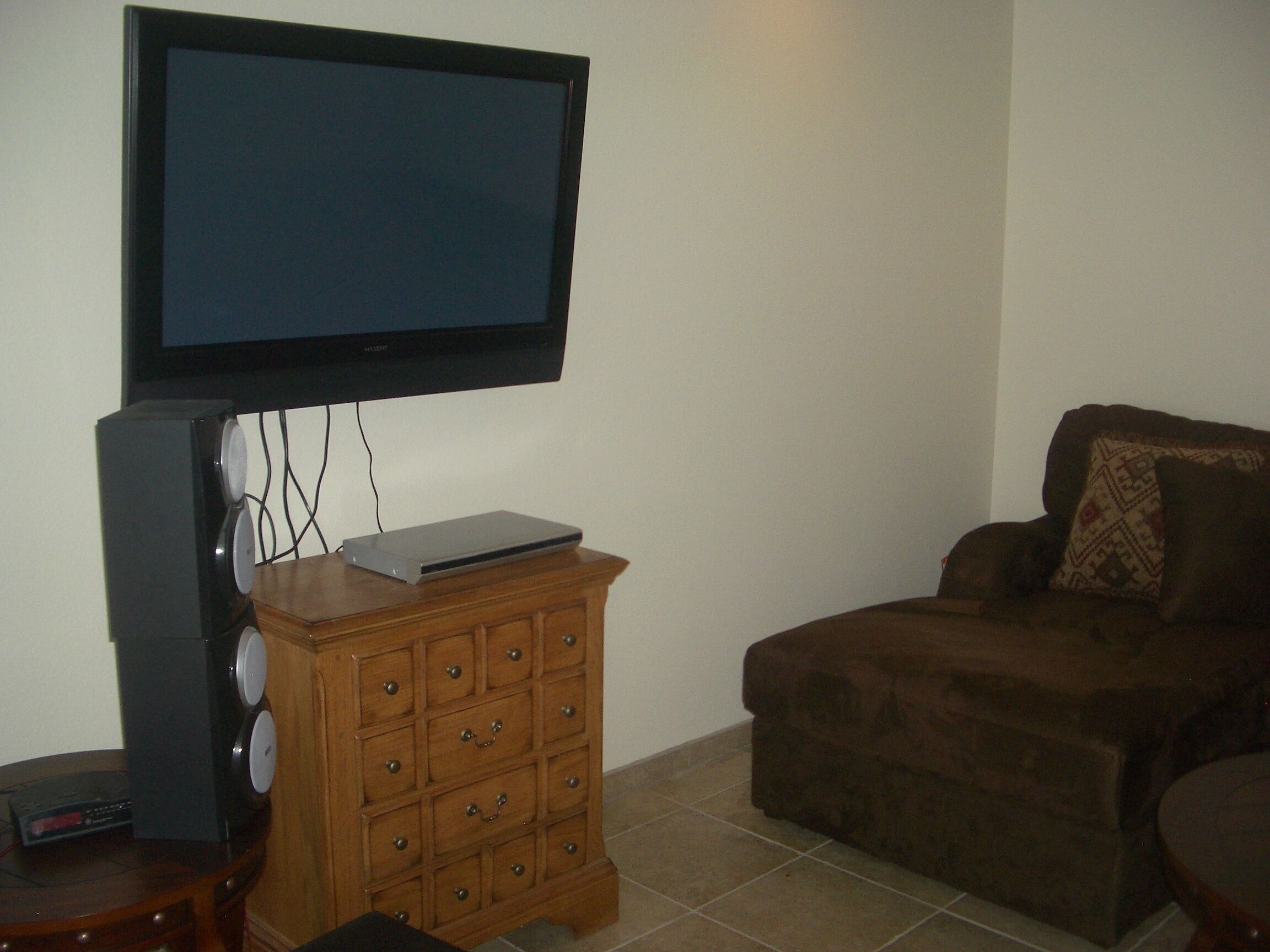 Een woonkamer met een flatscreen televisie aan de muur, een houten kast met lades, een bruine bank en een bijzettafeltje.