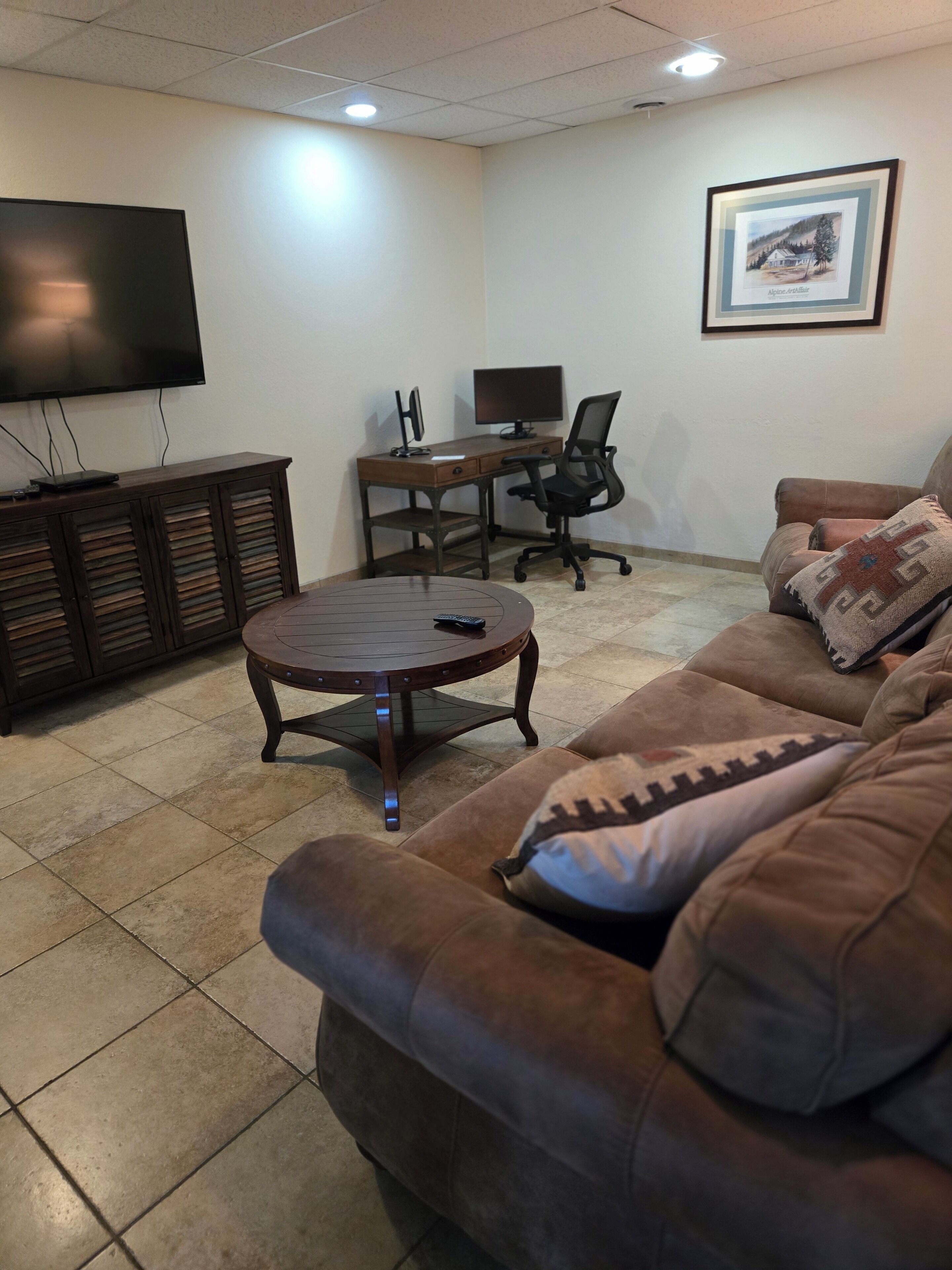 A furnished living room with a sofa, a round coffee table, a TV, a desk with a computer, and a framed picture on the wall.