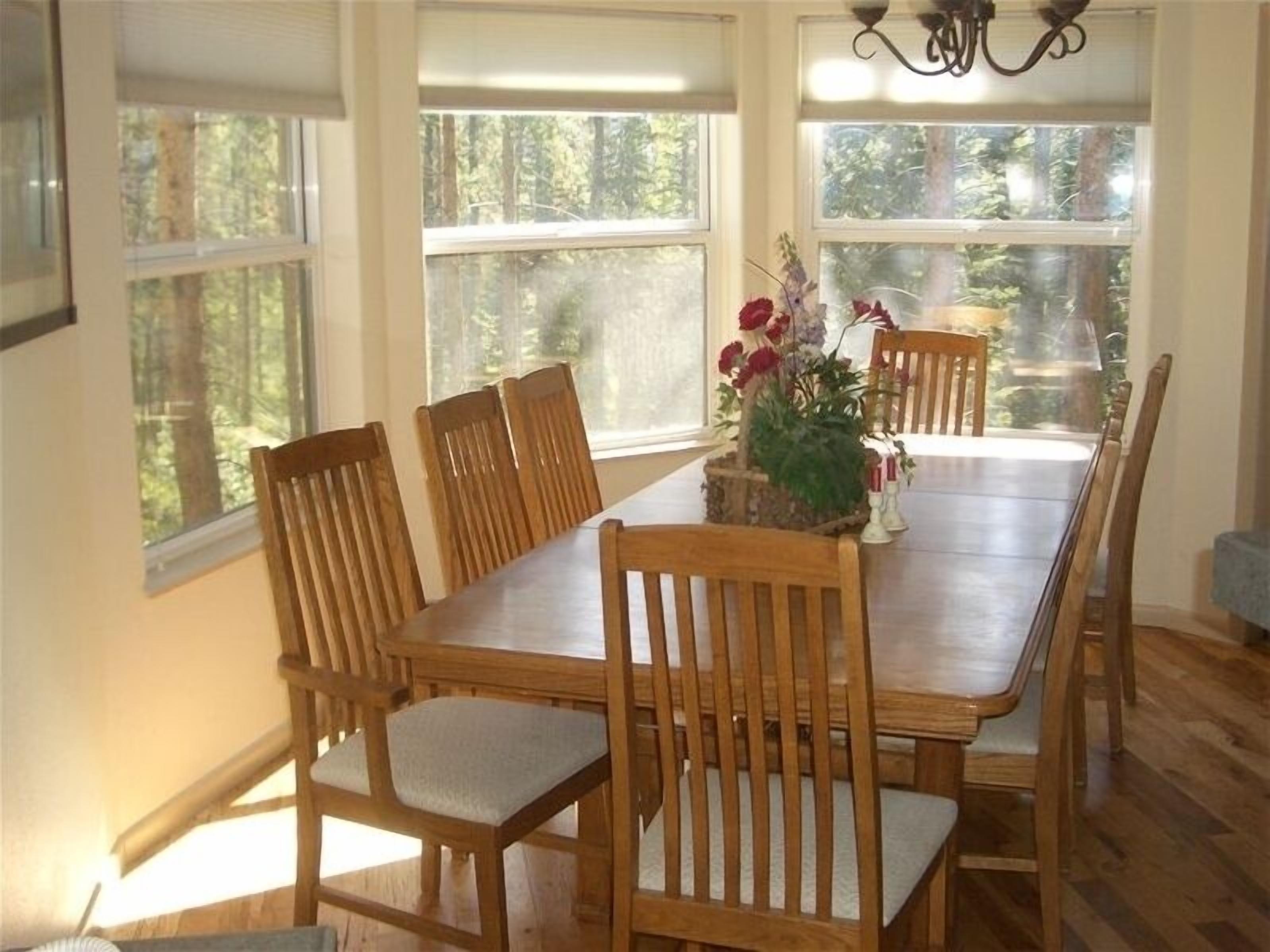 A dining room with a wooden table and chairs, large windows with a view of trees, and a chandelier.
