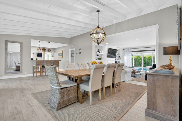 A spacious dining area with a large wooden table, cushioned chairs, and a hanging chandelier.