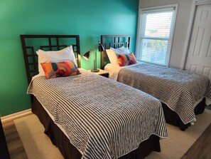 Une chambre avec deux lits, chacun doté de draps à rayures et d’une tête de lit au motif géométrique. Une table de chevet avec une lampe se trouve entre les deux lits, et une fenêtre avec des stores est située sur le côté droit.
