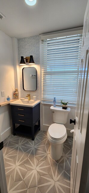Une salle de bain avec des toilettes blanches, un meuble-lavabo foncé doté de tiroirs, un miroir et une fenêtre avec des stores.