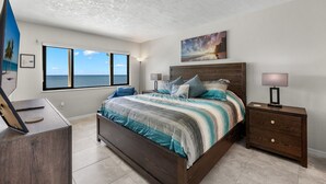 A bedroom with a large bed, a nightstand, a desk, and a window with a view of the ocean. - Riviera Club 9th floor Direct Beach Front IAN UPDATED AND READY!!! (Fort Myers Beach)