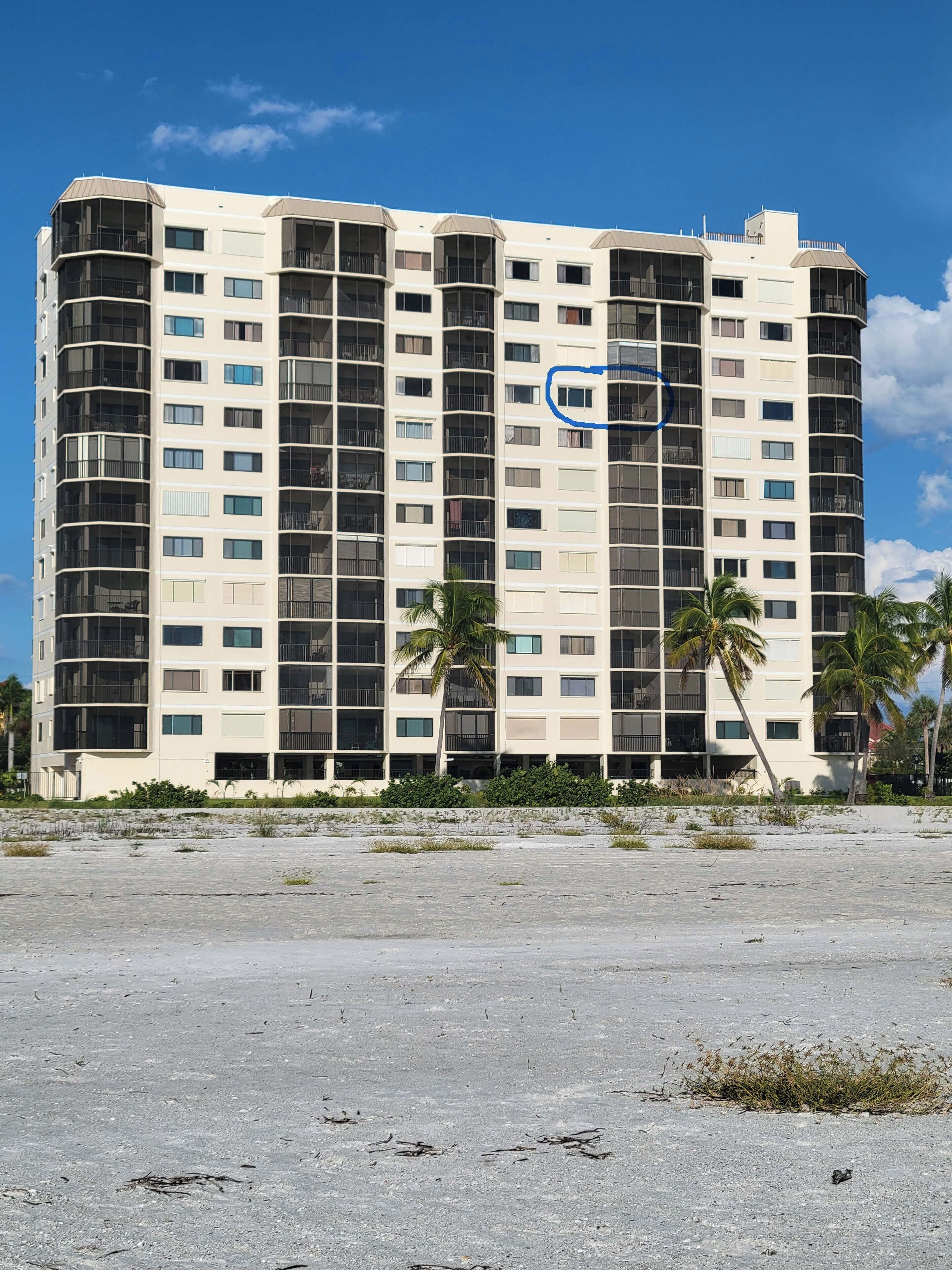 A multi-story hotel with balconies, palm trees, and a sandy foreground.