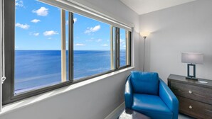 A room with a large window overlooking the sea, a blue chair, and a nightstand with a lamp. - Riviera Club 9th floor Direct Beach Front IAN UPDATED AND READY!!! (Fort Myers Beach)
