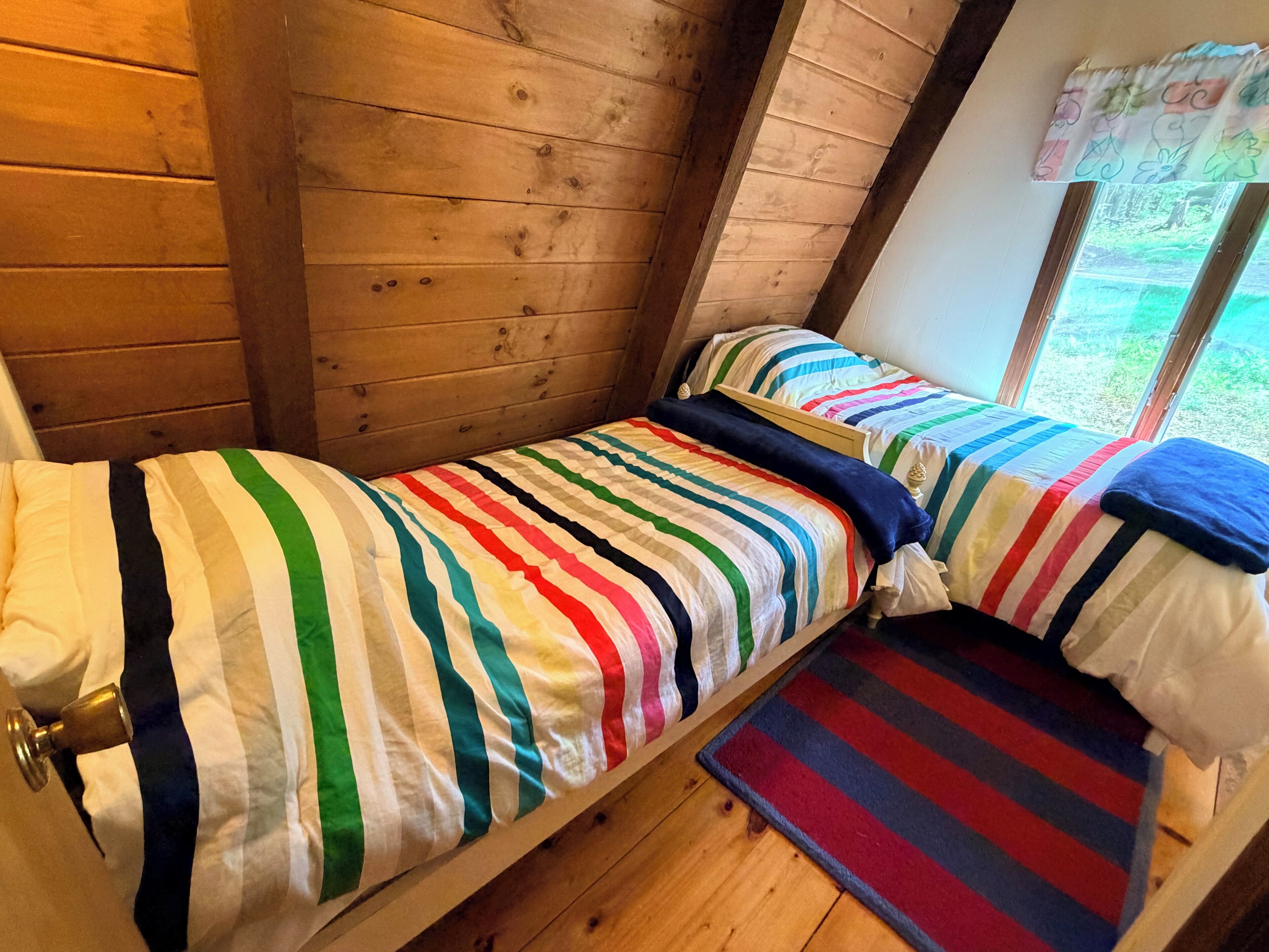 A single bed with a striped bedspread and pillows, a wooden floor, and a window with a view of greenery.