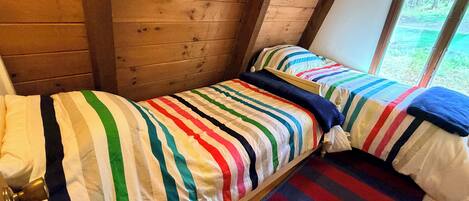 A single bed with a striped bedspread and pillows, a wooden floor, and a window with a view of greenery.