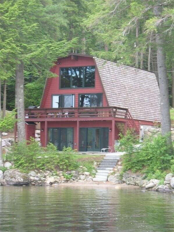 Situated by a body of water, this cabin features a steeply pitched roof, large windows, and a balcony area.