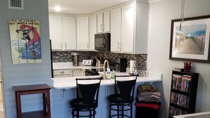 A kitchen with white cabinets, a black microwave, and a small island with two chairs. There is a wooden stool and a bookshelf with books. - Welcome to your affordable beach vacation (Gulf Shores)