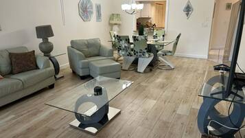 A modern living room with a glass coffee table, a sofa, a dining table with chairs, and a kitchen in the background.