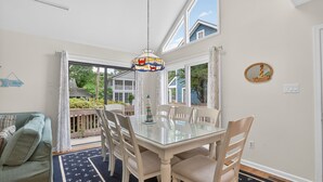 A dining area with a glass table, white chairs, a hanging light fixture, and a view of the outdoors.