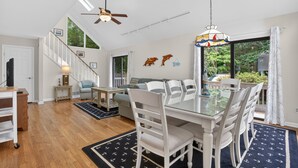 A spacious living area with a dining table, chairs, a sofa, a ceiling fan, and a view through large windows.