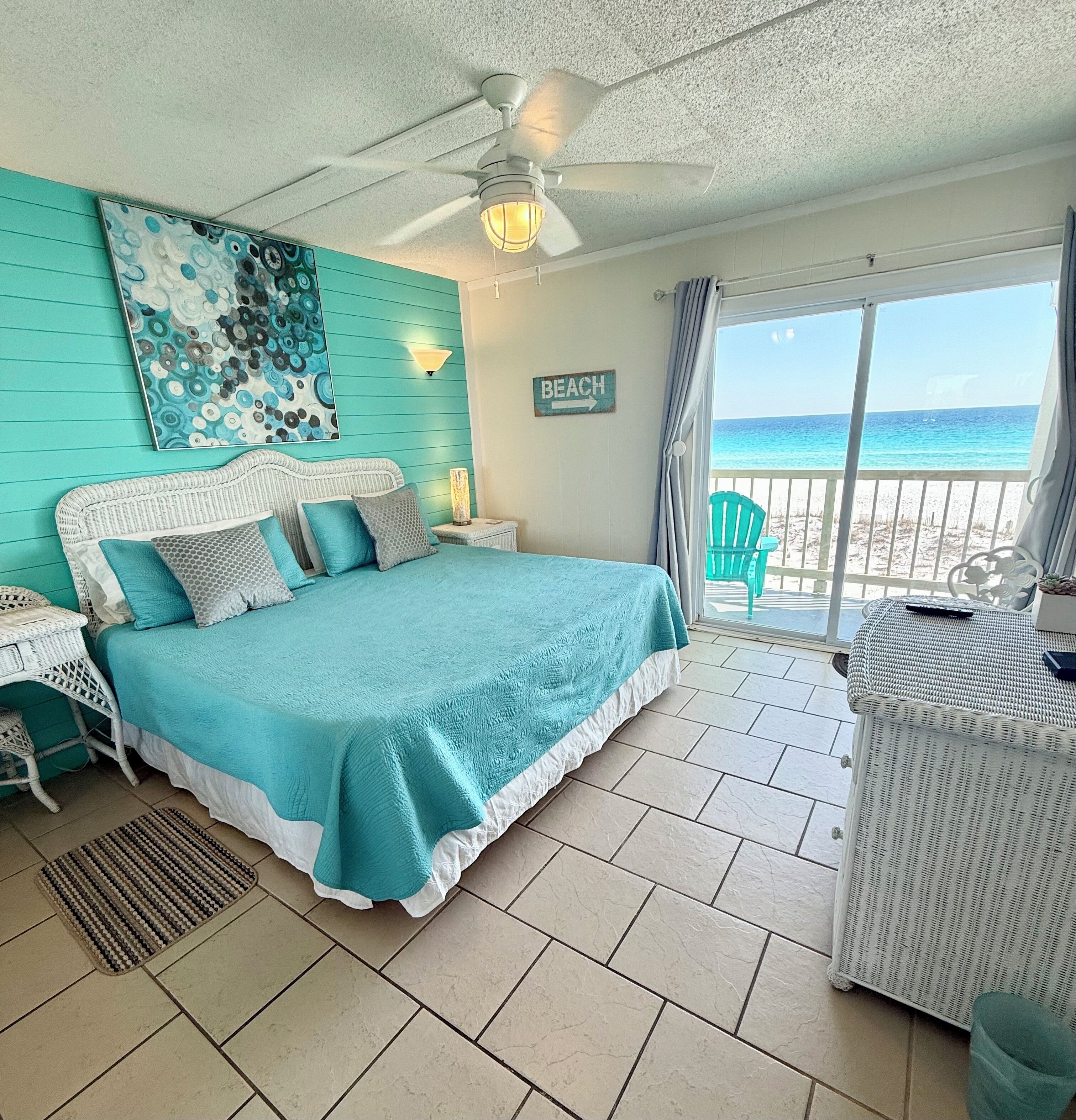 A bedroom with a bed, a nightstand, a lamp, a ceiling fan, a view of the beach, and a sign that says 'BEACH'.