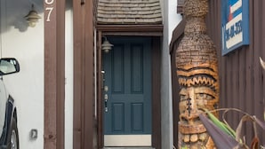 Un edificio con una puerta azul oscuro, un letrero de madera con el número '1037', un jarrón decorativo grande y una planta en maceta con follaje verde y flores rojas.