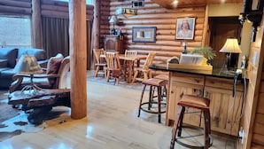 A rustic log cabin interior with a dining area, wooden chairs, a table, and a bar counter.