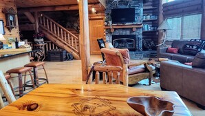 A log cabin interior with a wooden dining table, a fireplace, a stone wall, and a staircase leading to a loft area.