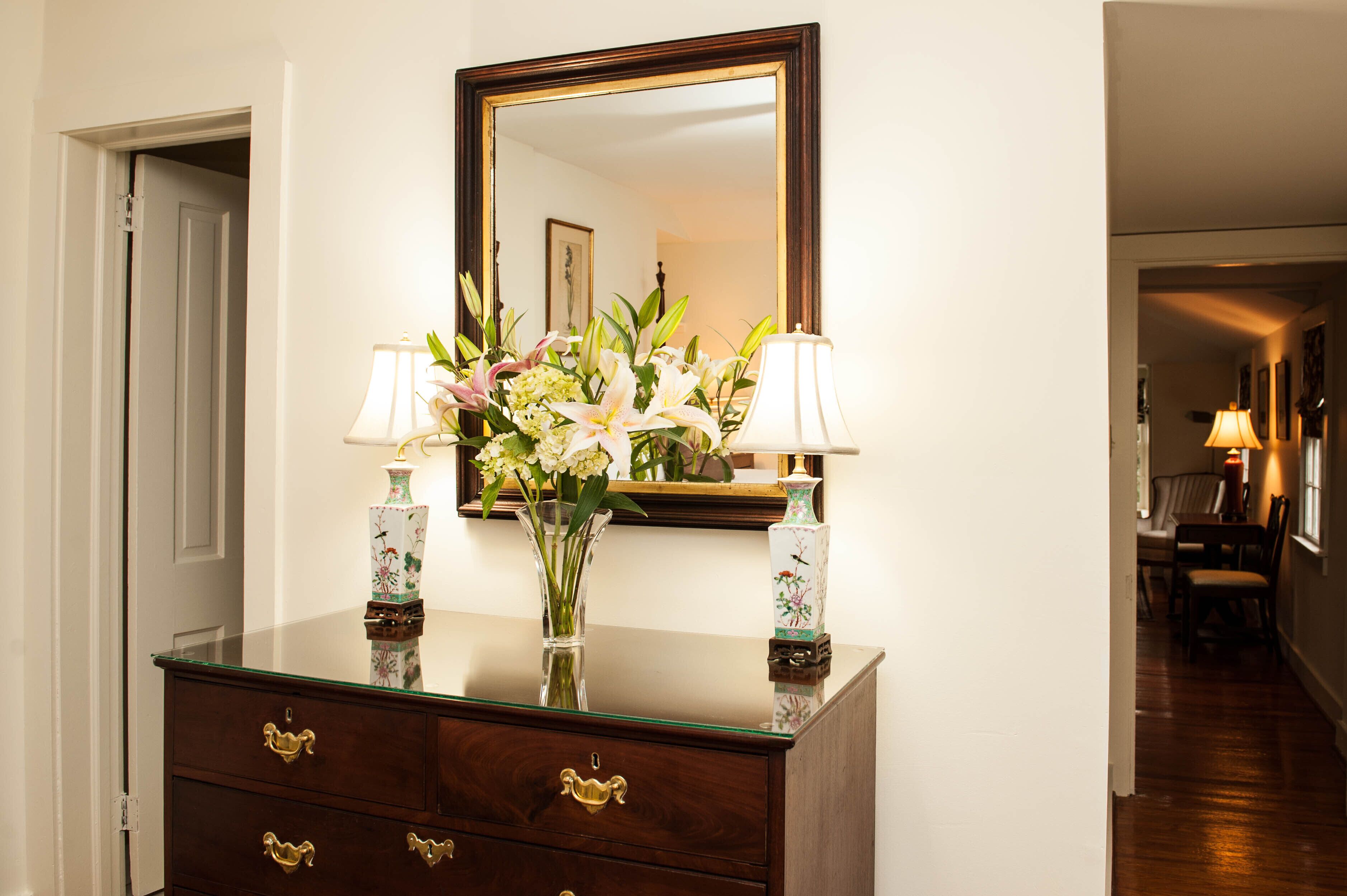 A wooden dresser with two lamps and a vase of flowers in front of a mirror.