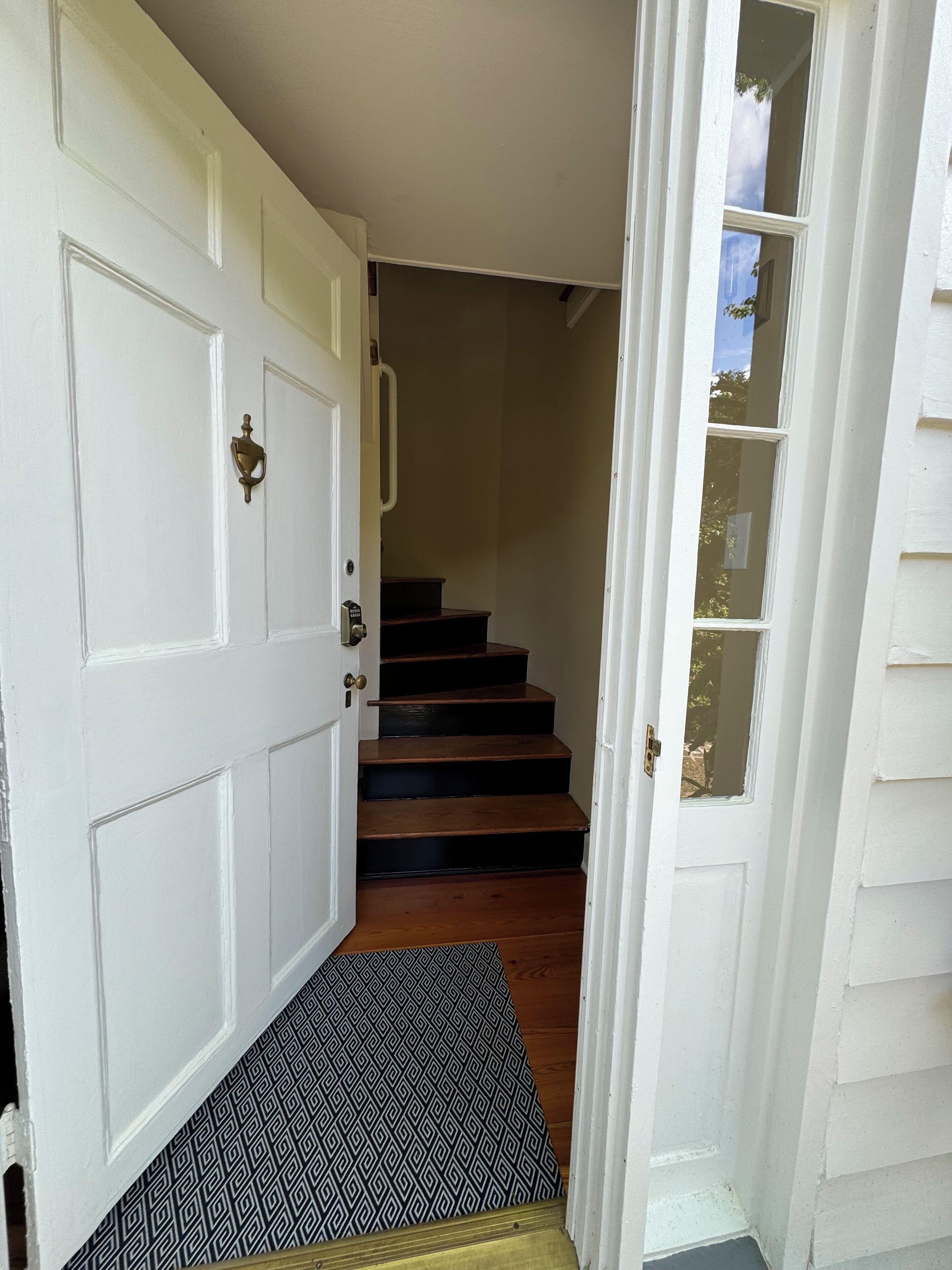 A white door with a brass handle, a black and white patterned doormat, and a set of wooden stairs leading to a landing.