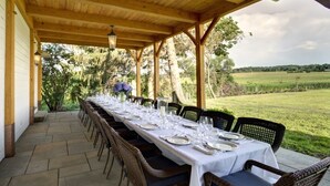 A covered outdoor dining area with a long table set for a meal, surrounded by wicker chairs, and overlooking a grassy field.