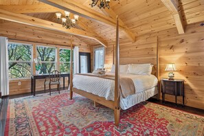 A four-poster bed with a quilted bedspread, a wooden desk with a chair, a nightstand with a lamp, and a large window with curtains.