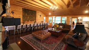 A spacious wooden interior with a dining area, a living room with a sofa and ottoman, and a large window offering a view.