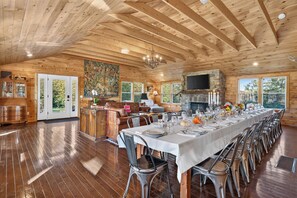 A spacious room with a long dining table set for a meal, wooden beams on the ceiling, a stone fireplace, and a large window offering a view of the outdoors.