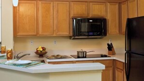Une cuisine avec des meubles de rangement en bois, un four à micro-ondes, un réfrigérateur et un plan de travail sur lequel se trouvent un panier de fruits et une bouilloire.