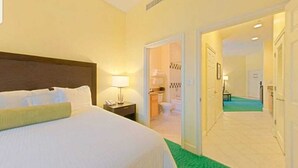 Une chambre d’hôtel avec un lit, une table de chevet, un ventilateur de plafond et une salle de bain visible à travers une porte ouverte.