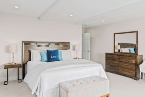 Quarto com cabeceira de madeira, cama com roupa de cama branca, um banco aos pés da cama, cômoda e espelho.