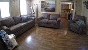 Una sala de estar con piso de madera, un sofá de cuero marrón, un sillón de cuero marrón y una mesa de centro de madera. Hay un cuadro en la pared y una cocina al fondo.