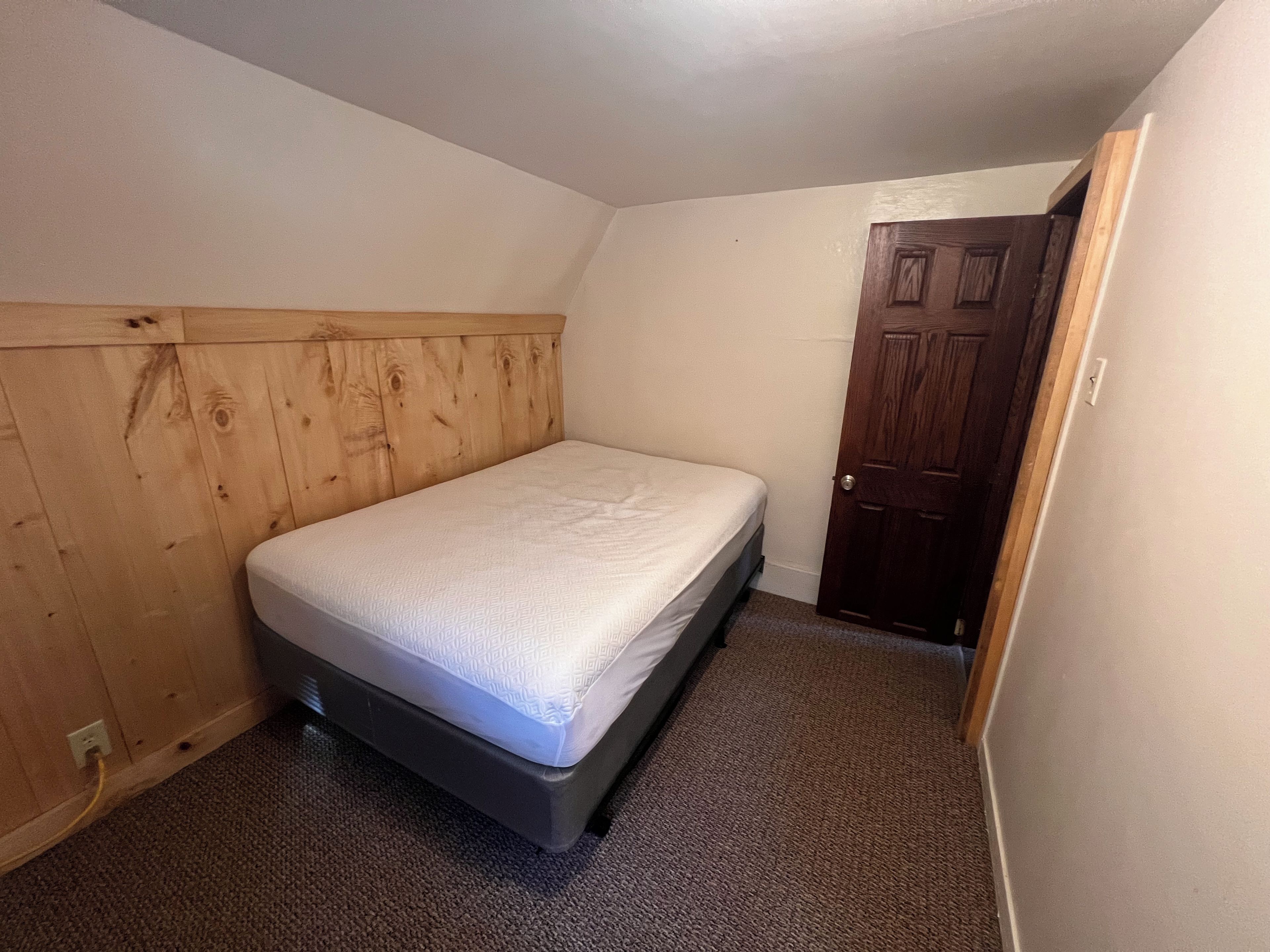 A small, single-bed room with a wooden headboard, carpeted floor, and a door.