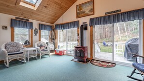 A cozy cabin interior with wicker furniture, a wood stove, and large windows overlooking a deck.